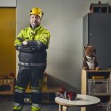 Perry Warren seisoo työhaalareissa ja kypärä päässä päiväkodin leikkitilassa, jossa on paljon leluja lattialla ja hyllyillä.